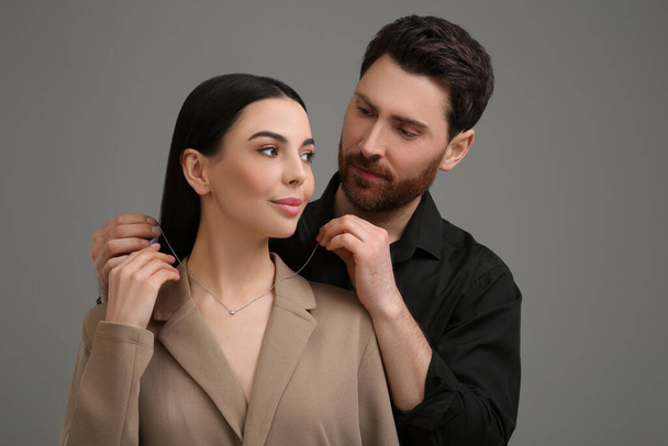 Man putting elegant necklace on beautiful woman against dark grey background - Фото, зображення