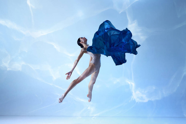 Light as feather. Shot of beautiful jumping ballerina with silk fabric over blue background. Ballet with silk. Concept of human emotions, art, dance, theater. Contemporary ballet - 写真・画像