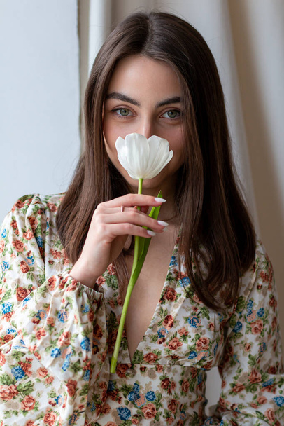 Pretty young woman in dress with flowers and dark hair with a tulip next to the window - Foto, Imagem