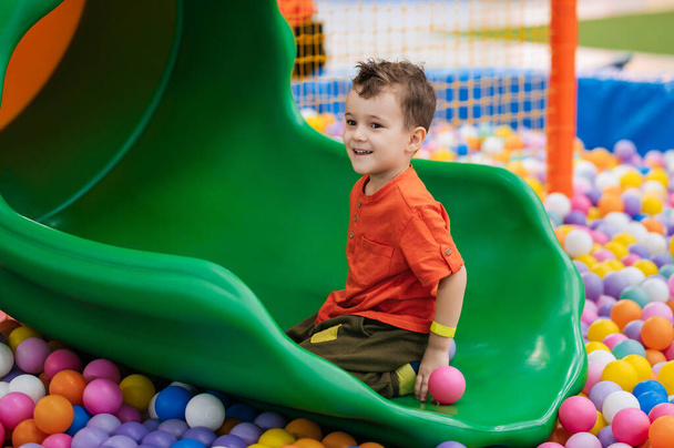 Счастливый мальчик играет в сухом бассейне с красочными мячами и скачет в них по горке. Развлекательный центр для детей. Активный отдых - Фото, изображение