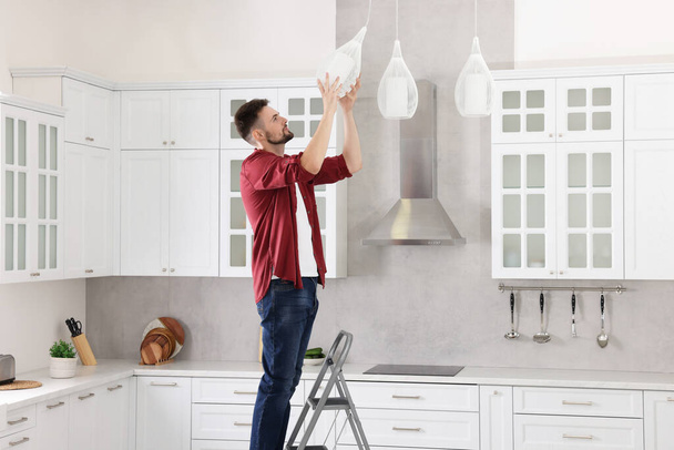 Young man installing ceiling lamp on stepladder in kitchen - Фото, зображення