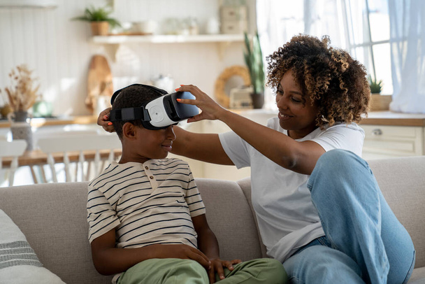 Mother limiting time child playing in virtual reality. Smiling African American mom sitting with child son on sofa taking off his vr glasses, putting kid in immersive world. Parenting in digital age - Fotó, kép