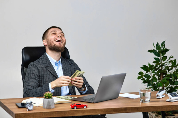 young bearded businessman working on laptop and counting dollars while sitting on chair at desk. - Photo, image