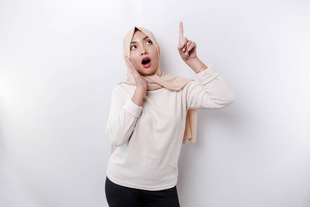 Shocked Asian Muslim woman wearing a headscarf pointing at the copy space on top of her, isolated by a white background - Zdjęcie, obraz