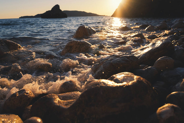 Скалистый галечный пляж с камнями на закате, вода с брызгами падает на берег - Фото, изображение
