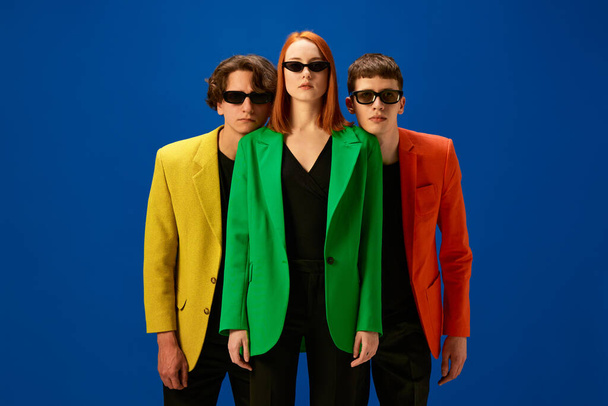 Young people wearing colorful jackets and dark sunglasses standing serious facial expression over blue background with. Concept of fashion, models, style - Photo, Image