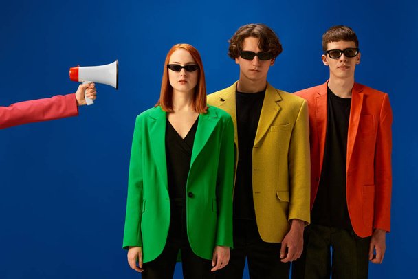 News, information. Portrait of young people wearing colorful jackets standing near loudspeaker over blue background. Concept of fashion, models, style - Φωτογραφία, εικόνα
