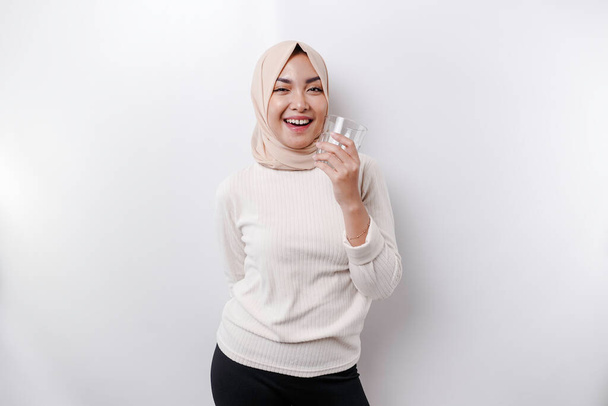 Joyful Asian Muslim woman wearing headscarf is drinking a glass of water, isolated on white background. - Φωτογραφία, εικόνα