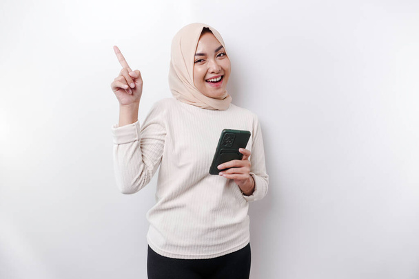 Excited Asian woman wearing hijab pointing at the copy space on top of her while holding her phone, isolated by white background - Foto, immagini