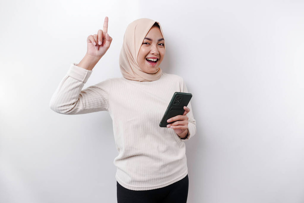 Excited Asian woman wearing hijab pointing at the copy space on top of her while holding her phone, isolated by white background - Фото, изображение