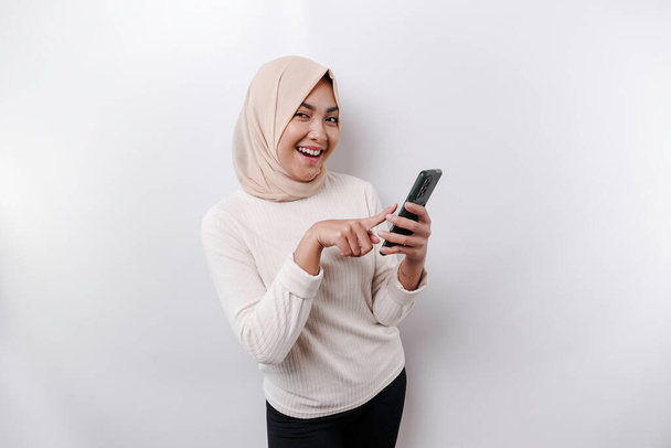 A happy Asian Muslim woman wearing a headscarf, holding her phone, isolated by white background - Fotó, kép