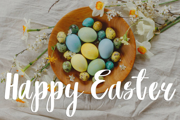 Happy Easter text sign on stylish πασχαλινά αυγά και ανθισμένα ανοιξιάτικα λουλούδια σε ξύλινο μπολ στο ρουστίκ τραπέζι. Σύγχρονη ευχετήρια κάρτα Πάσχα. Χειρόγραφα γράμματα - Φωτογραφία, εικόνα