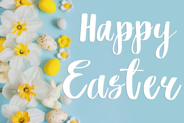 Joyeux signe de texte de Pâques sur des œufs de Pâques élégants, des fleurs de lapin et de jonquilles jaunes à plat sur fond bleu. Carte de vœux de Pâques moderne. Lettrage manuscrit - Photo, image