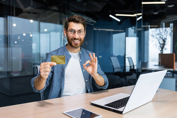 Successful businessman inside office building at workplace smiling and looking at camera, mature adult man holding and showing bank credit card, smiling contentedly. - Photo, Image