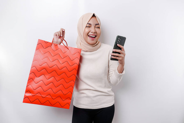 Portrait Asian Muslim woman standing excited holding an online shopping bag and her smartphone, studio shot isolated on white background - 写真・画像