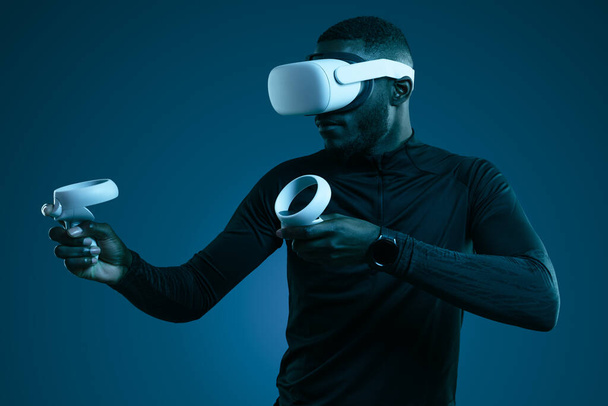 Bearded African American man in virtual reality goggles and black turtleneck standing with raised arms, and holding controllers while playing videogame in dark room with neon light - Foto, afbeelding