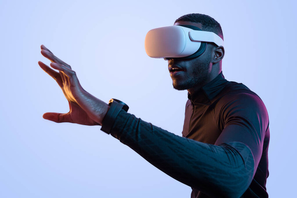Side view of concentrated young African American man with dark hair and beard in black athletic clothes, and VR glasses touching air while exploring virtual reality in neon studio - Foto, imagen