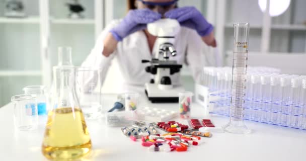 Flask con liquido giallo e mucchio di pillole sparse sul tavolo contro donna esaminando campione al microscopio. Medicinali e olio in laboratorio - Filmati, video