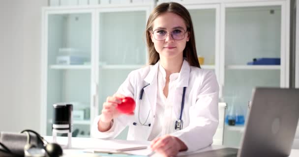 Θετική γυναίκα σε γυαλιά κρατά κόκκινο παιχνίδι καρδιά στο χέρι κοιτάζοντας στην κάμερα και ελαφρώς χαμογελώντας. Γιατρός με ιατρική στολή κάθεται στο τραπέζι σε αργή κίνηση γραφείου - Πλάνα, βίντεο
