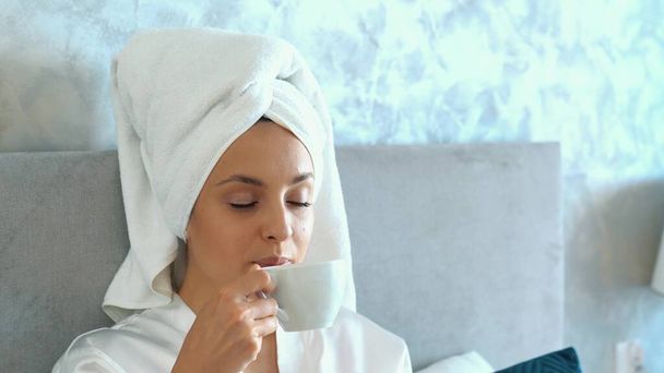 Pretty happy woman in bath robe and with towel on head lying on bed after shower drinking coffee enjoying time for relax. Beautiful Caucasian female resting on bed in bedroom at cozy home. Slow motion - Fotó, kép