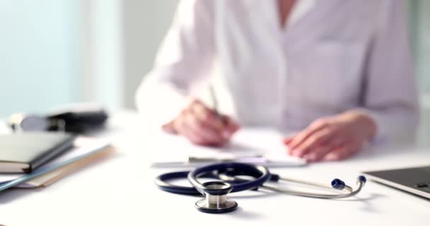 Stethoscope lies on white table against doctor writing on clipboard near laptop. Professional medical equipment in modern clinic office - Footage, Video