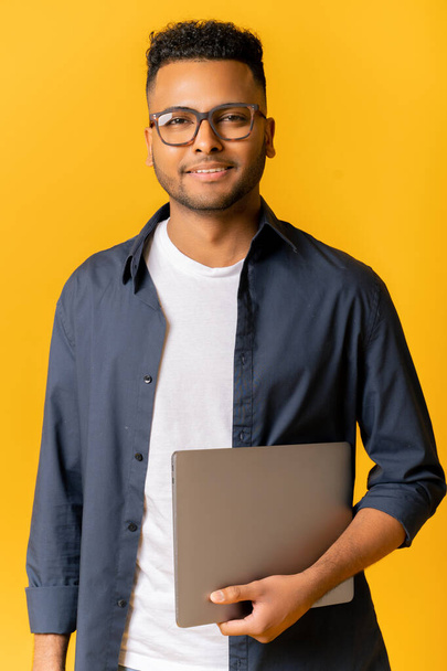Portrait of successful indian man in glasses standing and carrying laptop, looking at the camera isolated on yellow, cheerful male entrepreneur, freelancer or student smiling - Foto, imagen