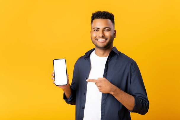 Happy excited young indian man looking at camera pointing finger at empty phone screen, paying attention at copy space, advertising new promotion, presenting sale offer standing isolated on yellow - Valokuva, kuva