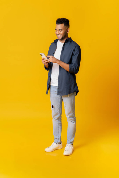 Online kommunikáció. Teljes hosszúságú portré indiai férfi használ okostelefon álló elszigetelt sárga, arab férfi diák srác görgetés, ellenőrzése értesítések, dolgozik a távolságot, SMS - Fotó, kép