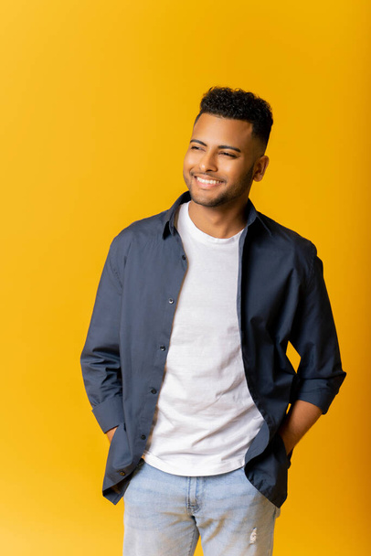 Portrait of carefree indian guy in casual shirt standing isolated on yellow background, arab man posing in studio, holding hands in pockets of jeans, relaxed serene handsome man looking away - Foto, afbeelding
