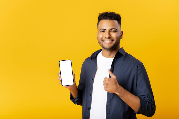 Happy young indian man looking at camera, holding smartphone with empty phone screen and showing thumb up in sign of approving, advertising new promotion, recommending new app isolated on yellow - Foto, imagen