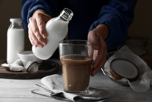 Woman holding bottle with coconut milk near glass of coffee at white wooden table, closeup - Fotografie, Obrázek