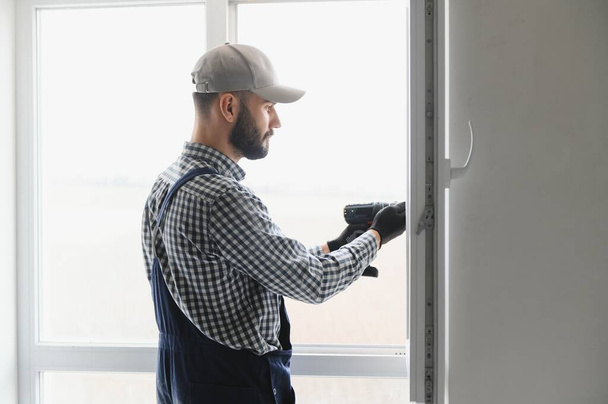 Workman in overalls installing or adjusting plastic windows in the living room at home. - Foto, Imagen