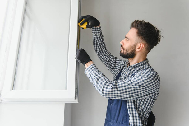 Workman in overalls installing or adjusting plastic windows in the living room at home. - Φωτογραφία, εικόνα