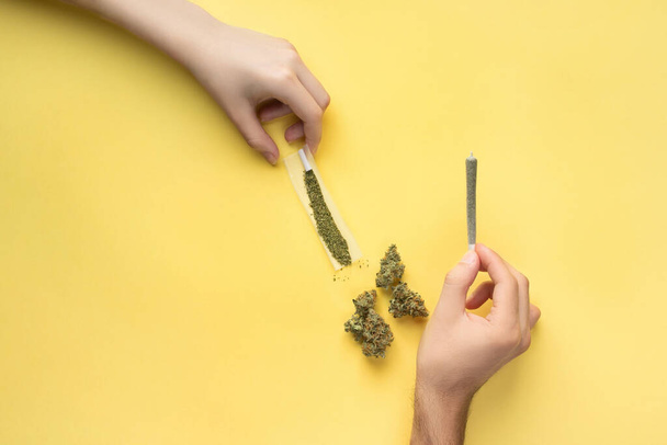 Las articulaciones con marihuana medicinal en manos de una chica y un chico, junto a ellas están los brotes de cannabis, el papel y un filtro. Sobre un fondo amarillo brillante - Foto, Imagen