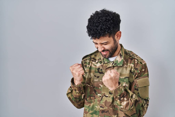 Арабский мужчина в камуфляжной армейской форме очень счастлив и взволнован, делая жест победителя с поднятыми руками, улыбаясь и крича об успехе. празднование концепции.  - Фото, изображение