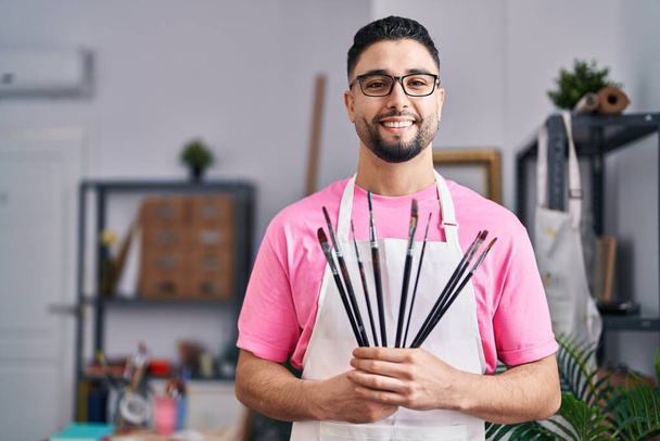 Giovane arabo uomo artista sorridente fiducioso tenendo pennelli allo studio d'arte - Foto, immagini