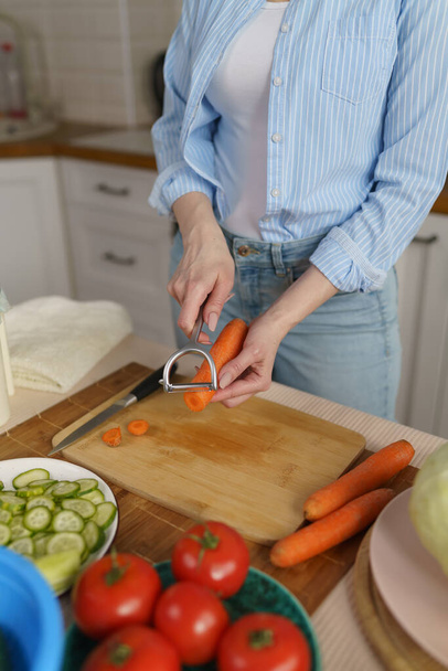 House wife prepares lunch with fresh vegetables. Woman peeling carrots with a peeler tool. Female person cooking healthy vegetarian meal at home - Fotografie, Obrázek