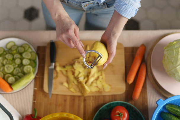 Woman peeling potato for dinner. Person cooking vegetables at home, shot from above. Female hands using food peeler tool - Foto, Bild