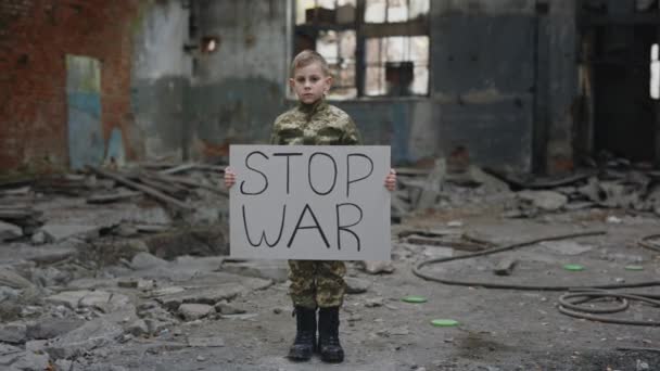 Retrato del niño pequeño con uniforme militar protestando por la guerra, sosteniendo pancartas con inscripción Stop War. Hombre Kid Activista mirando la cámara. Niño contra la guerra. Patriota. - Metraje, vídeo