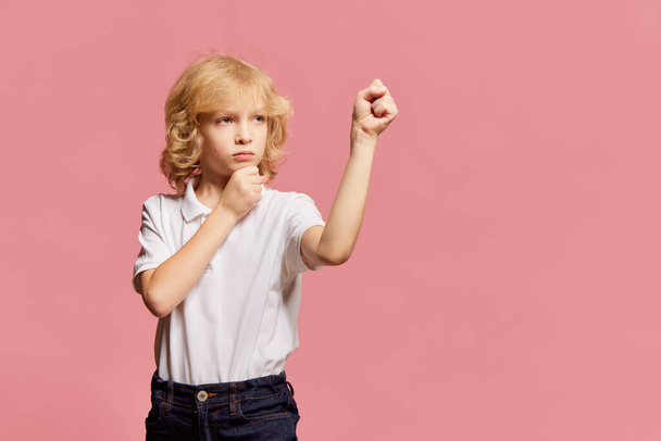 Game and imagination. Little boy, child posing with serious face, showing fists against pink studio background. Model in white shirt. Concept of childhood, emotions, facial expression. Ad - Foto, afbeelding