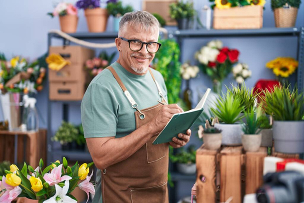 Middle age grey-haired man florist smiling confident writing on notebook at flower shop - Photo, image