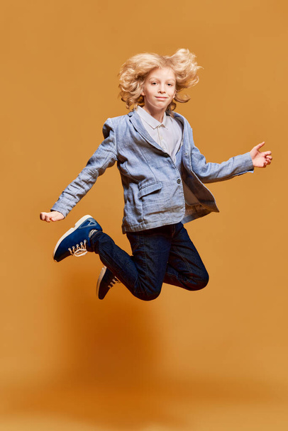 Full-length portrait of little boy, child in jacket jumping, posing against orange studio background. Model with blonde curly hair. Concept of childhood, emotions, facial expression. Ad - 写真・画像