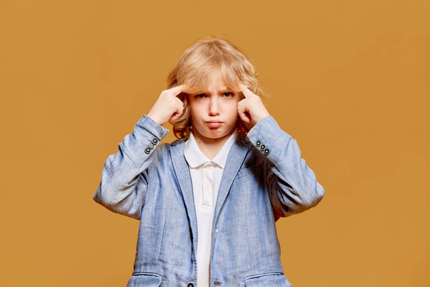 Little boy, child with curly blonde hair posing with fingers to head against orange studio background. Thinking process. Concept of childhood, emotions, facial expression. Ad - Φωτογραφία, εικόνα