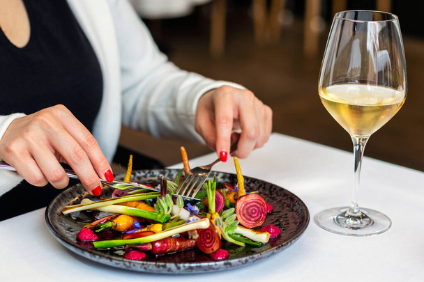 Κοντινές λεπτομέρειες της γυναίκας τρώει ορεκτικό φρέσκα ανάμεικτα λαχανικά σαλάτα σε γκουρμέ εστιατόριο. Λευκό ποτήρι κρασί στο τραπέζι δίπλα στο πιάτο. - Φωτογραφία, εικόνα