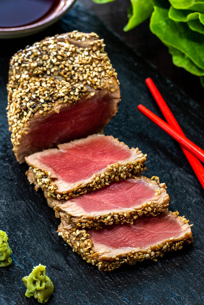 Vertical close up of appetizing sesame crusted tuna steak. Medium rare Yellow fin tuna slices on black slate with wasabi, soy sauce and chopsticks. - Photo, Image