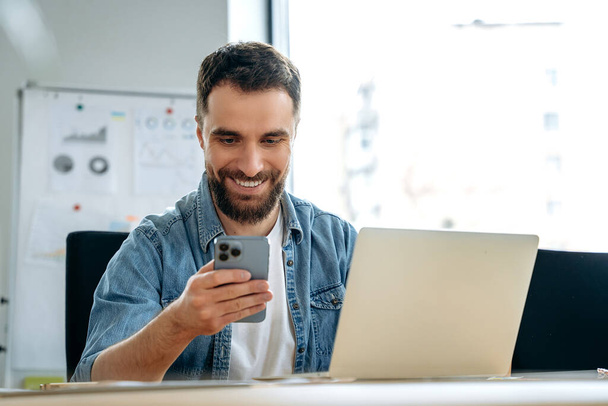 Удовлетворенный кавказский мужчина с бородой, фрилансер, программист, сео, сидит за столом в офисе, пользуется своим смартфоном, общается в социальных сетях, ведет рабочую переписку, смотрит новости, улыбается - Фото, изображение