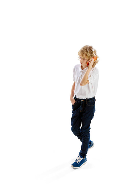 Isometric view. Little boy, child with blonde hair in shirt walkign and talking on mobile phone against white background. Concept of childhood, emotions, facial expression. Ad - Zdjęcie, obraz