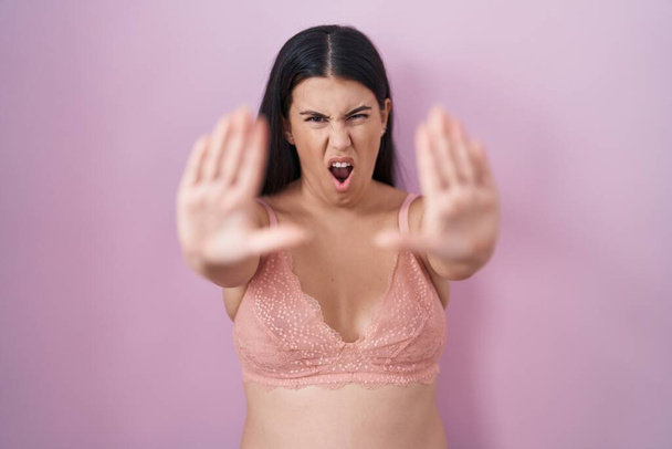 Jeune femme hispanique portant un soutien-gorge rose faisant geste d'arrêt avec les paumes des mains, expression de colère et de frustration  - Photo, image