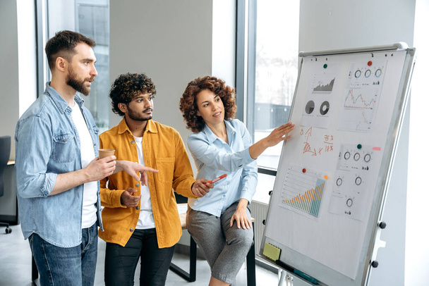 Teamwork. Successful multiracial colleagues, stand in the office near a marker white board, analyze the results of work, financial statistic, predict financial risks and profitability in a boardroom - 写真・画像