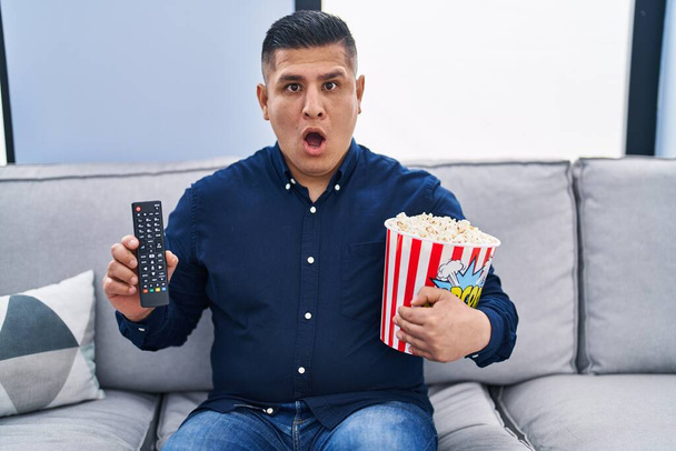 Hispanic young man eating popcorn using tv control in shock face, looking skeptical and sarcastic, surprised with open mouth  - Foto, imagen
